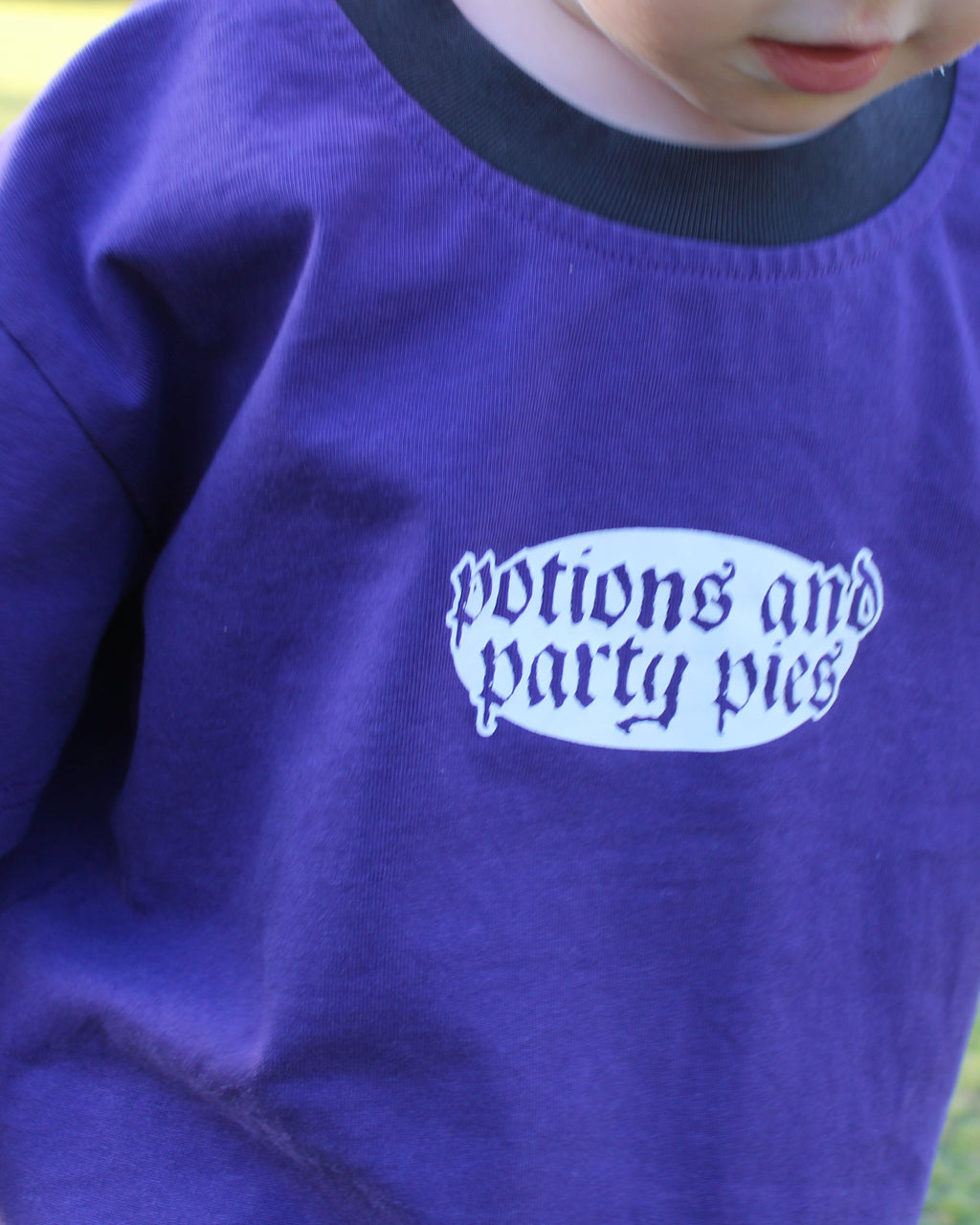 Child wearing a purple shirt with 'potions and party pies' text outdoors. Black collar.