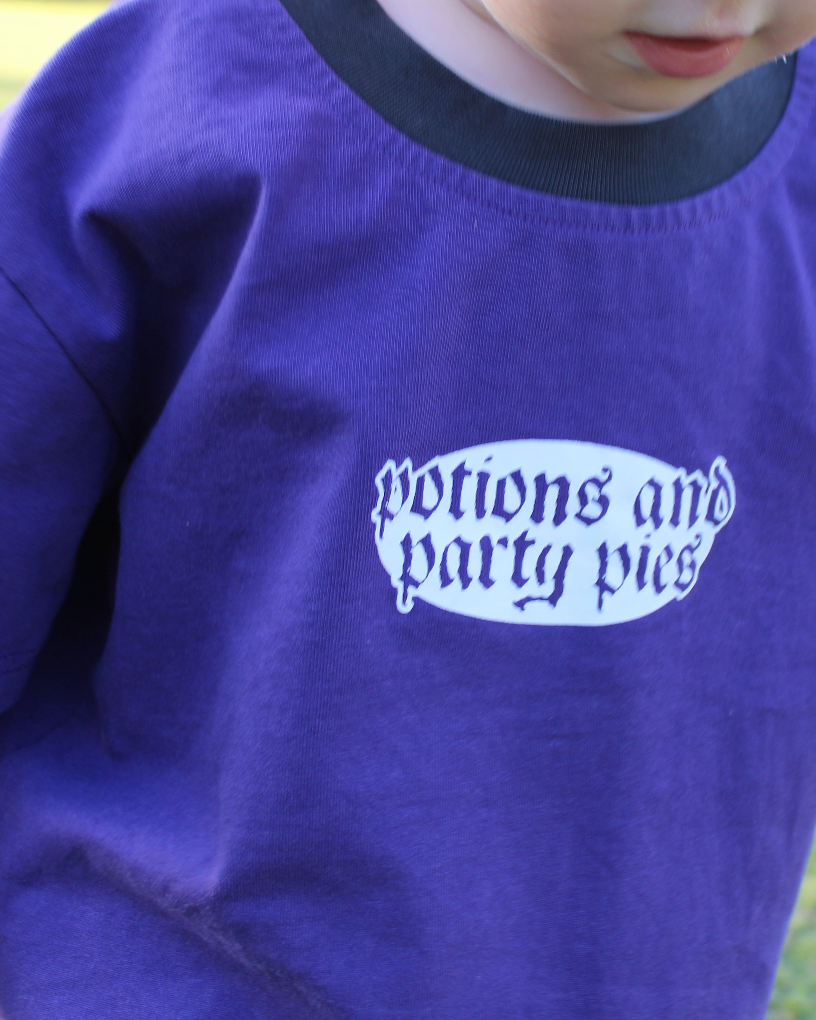 Child wearing a purple shirt with 'potions and party pies' text outdoors. Black collar.