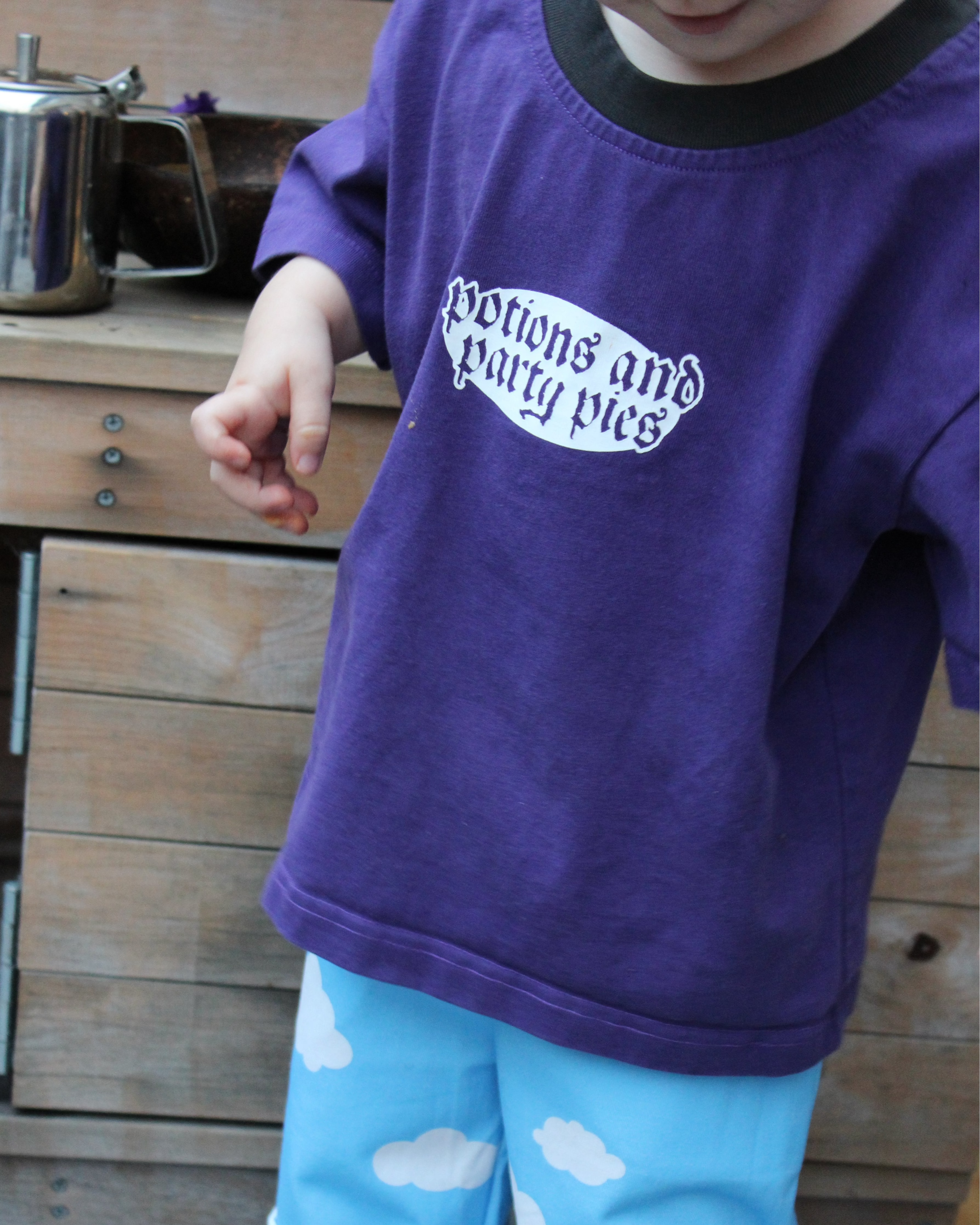Child wearing a purple shirt with text that reads 'potions and party pies' and blue pants with cloud patterns, standing in front of a wooden mud kitchen.