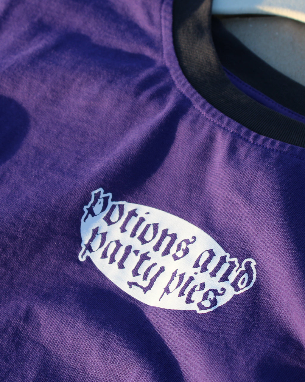Purple shirt with a black collar on a hanger. 'Potions and Party Pies' screenprint text on a plain background