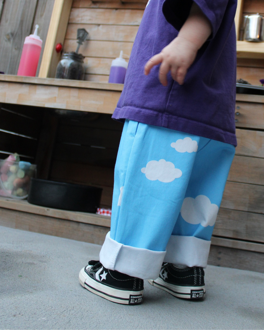 Child wearing a purple shirt and blue pants with cloud patterns standing in front of a wooden mud kitchen.