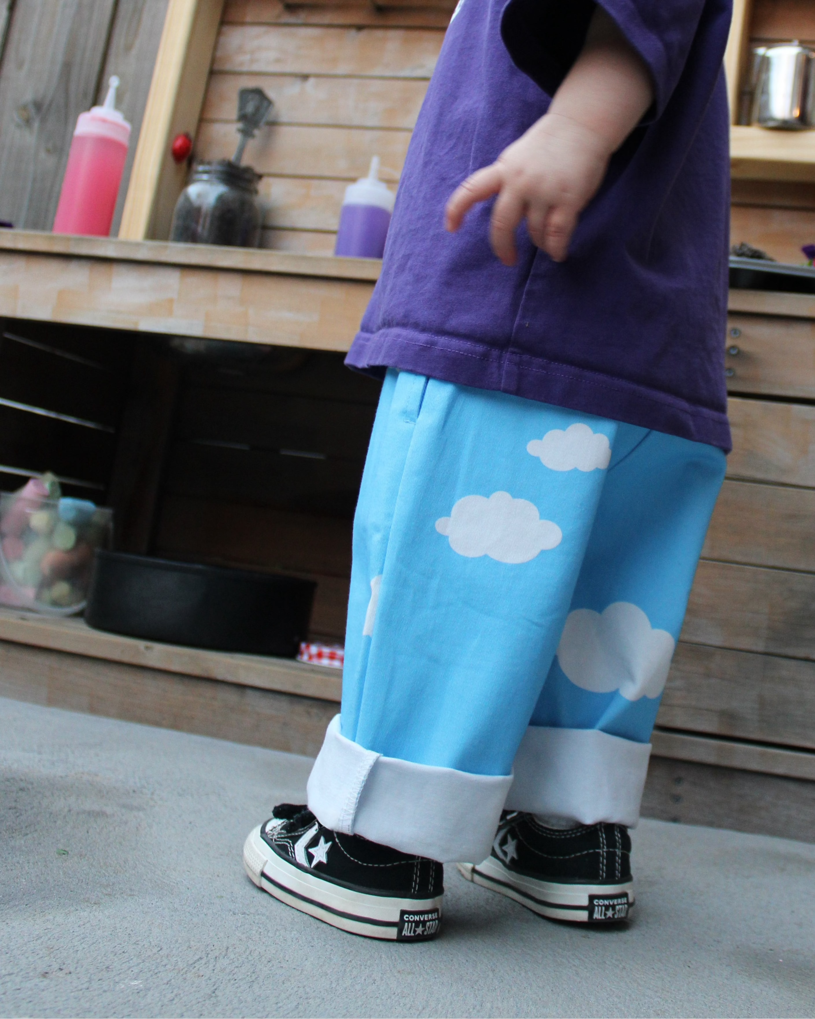 Child wearing a purple shirt and blue pants with cloud patterns standing in front of a wooden mud kitchen.
