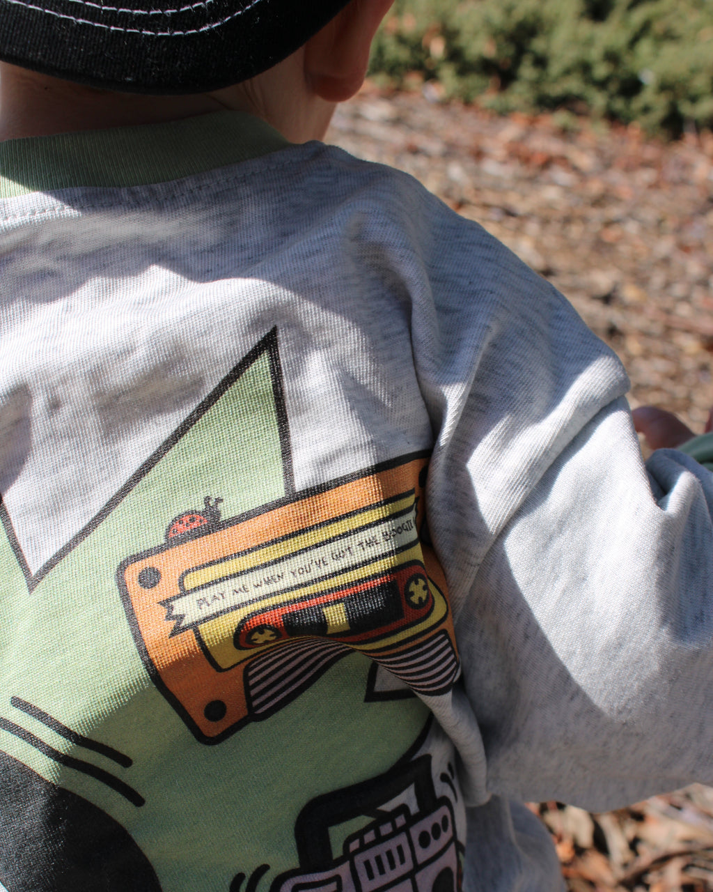 Child wearing a grey marle shirt with a colorful music-themed graphic design outdoors. Cassette reads 'play me when you've got the boogie bug'