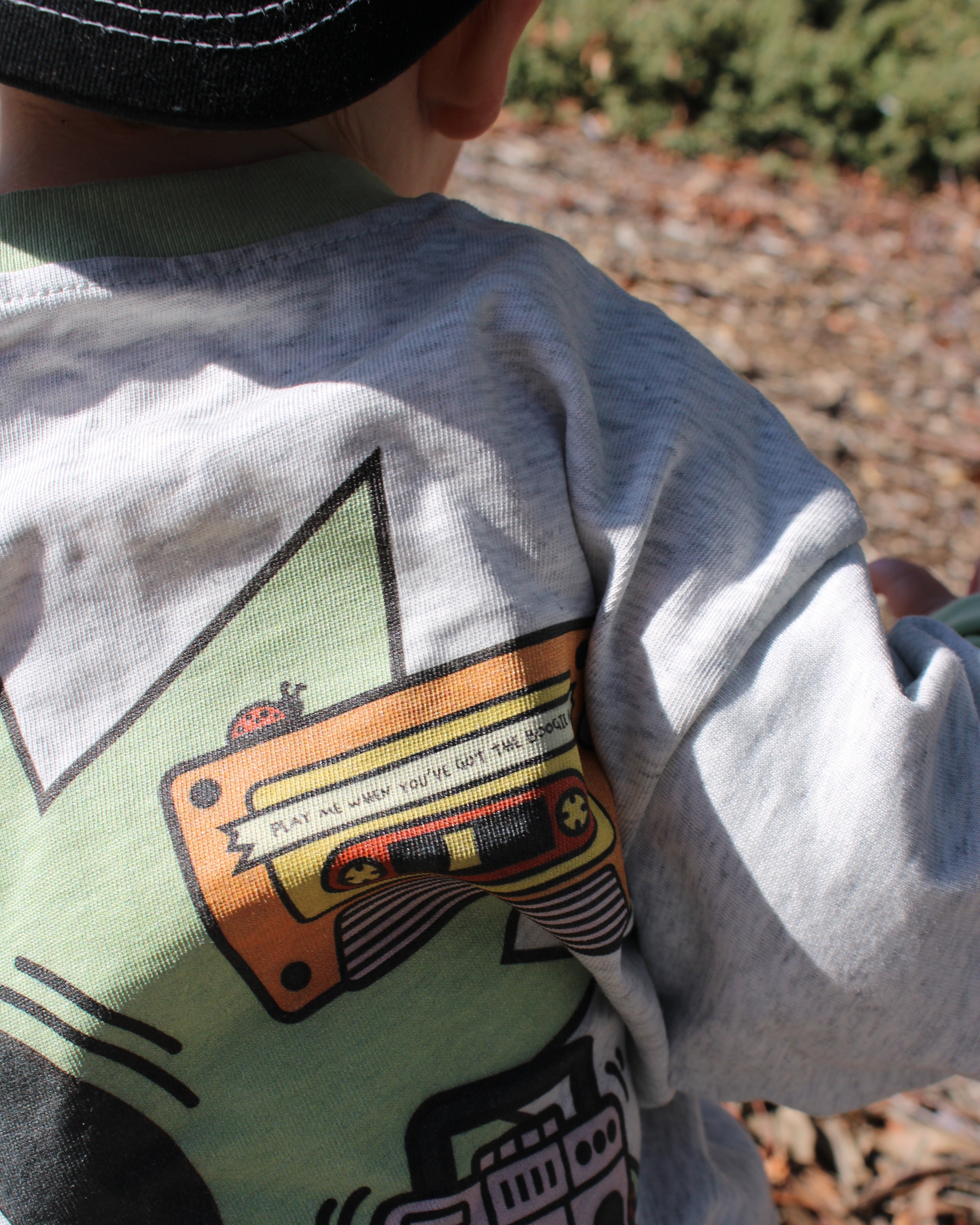 Child wearing a grey marle shirt with a colorful music-themed graphic design outdoors. Cassette reads 'play me when you've got the boogie bug'