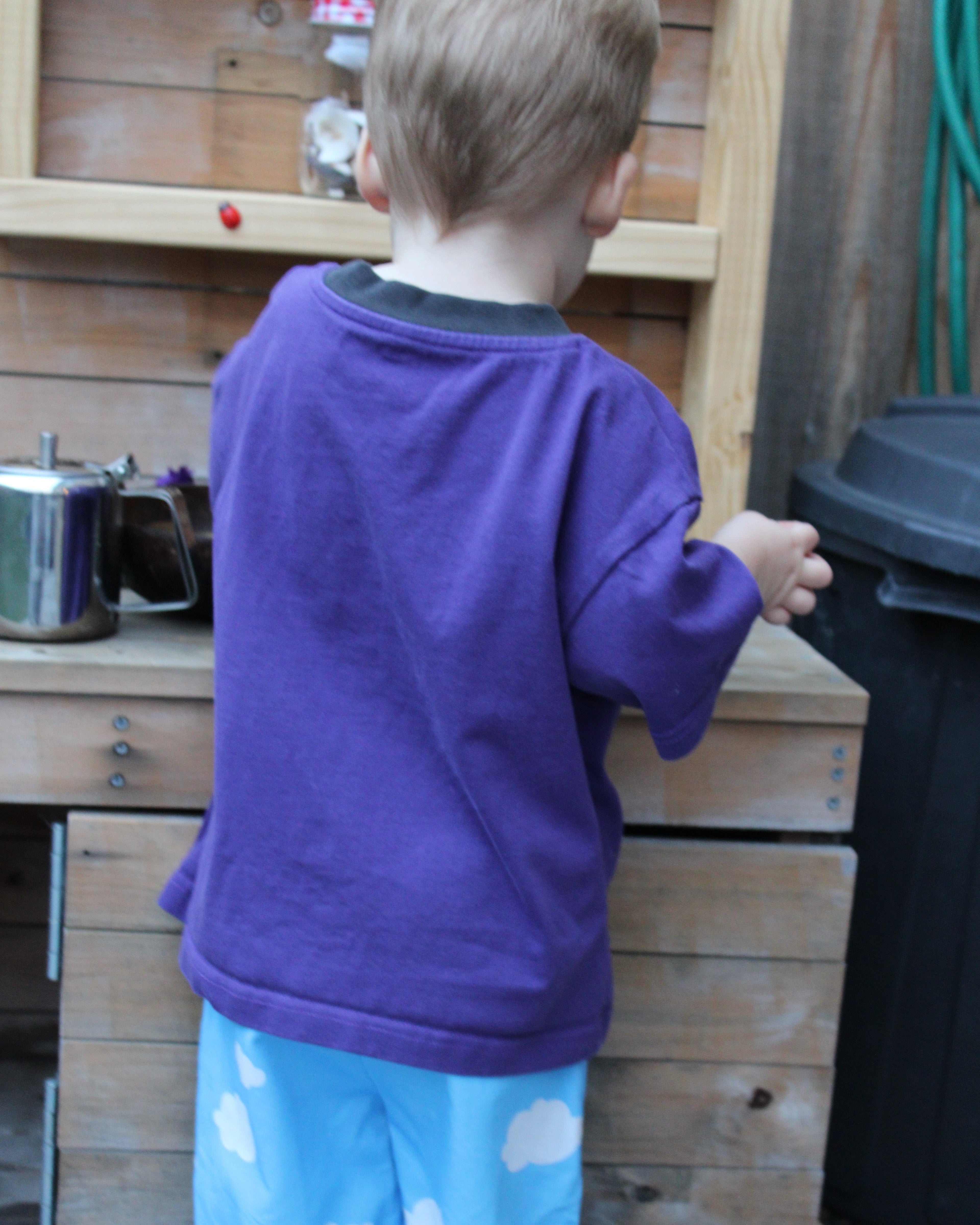 Child in a purple t-shirt and blue pants with cloud pattern standing next to a wooden play kitchen set.