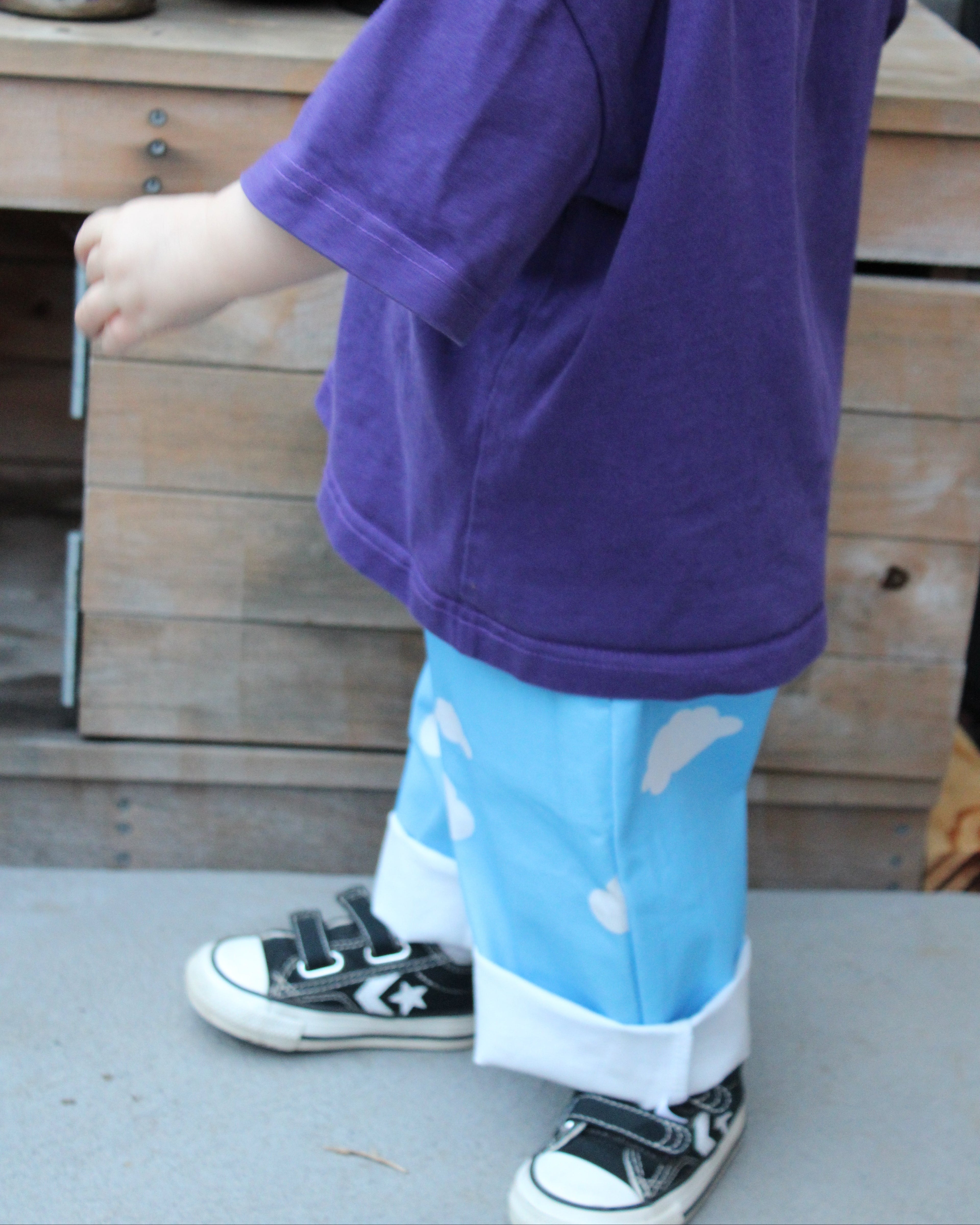 Child wearing a purple shirt and blue pants with cloud patterns, standing on a concrete surface.