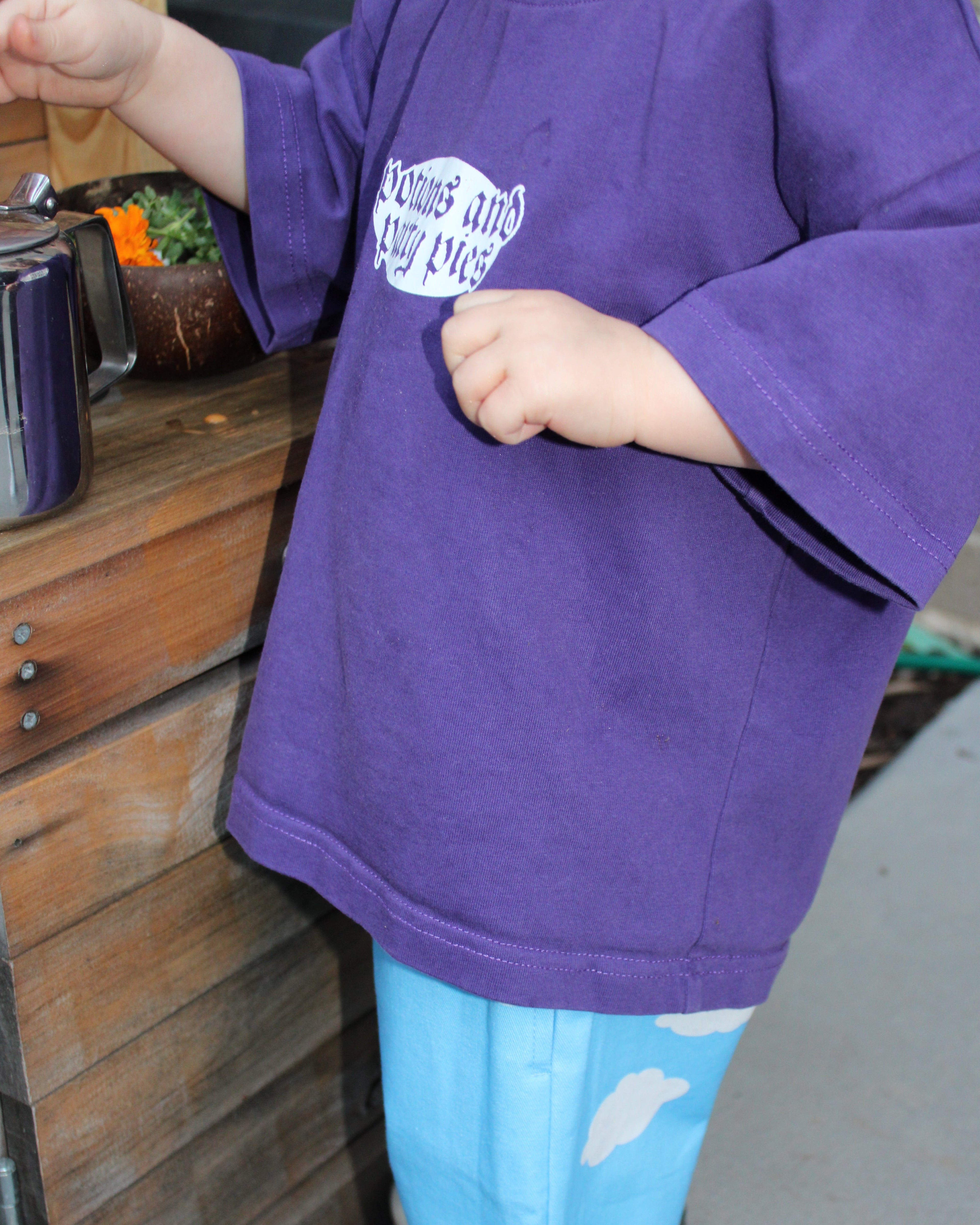 Child wearing a purple t-shirt and blue pants with cloud patterns in a mud kitchen, clenching fist.