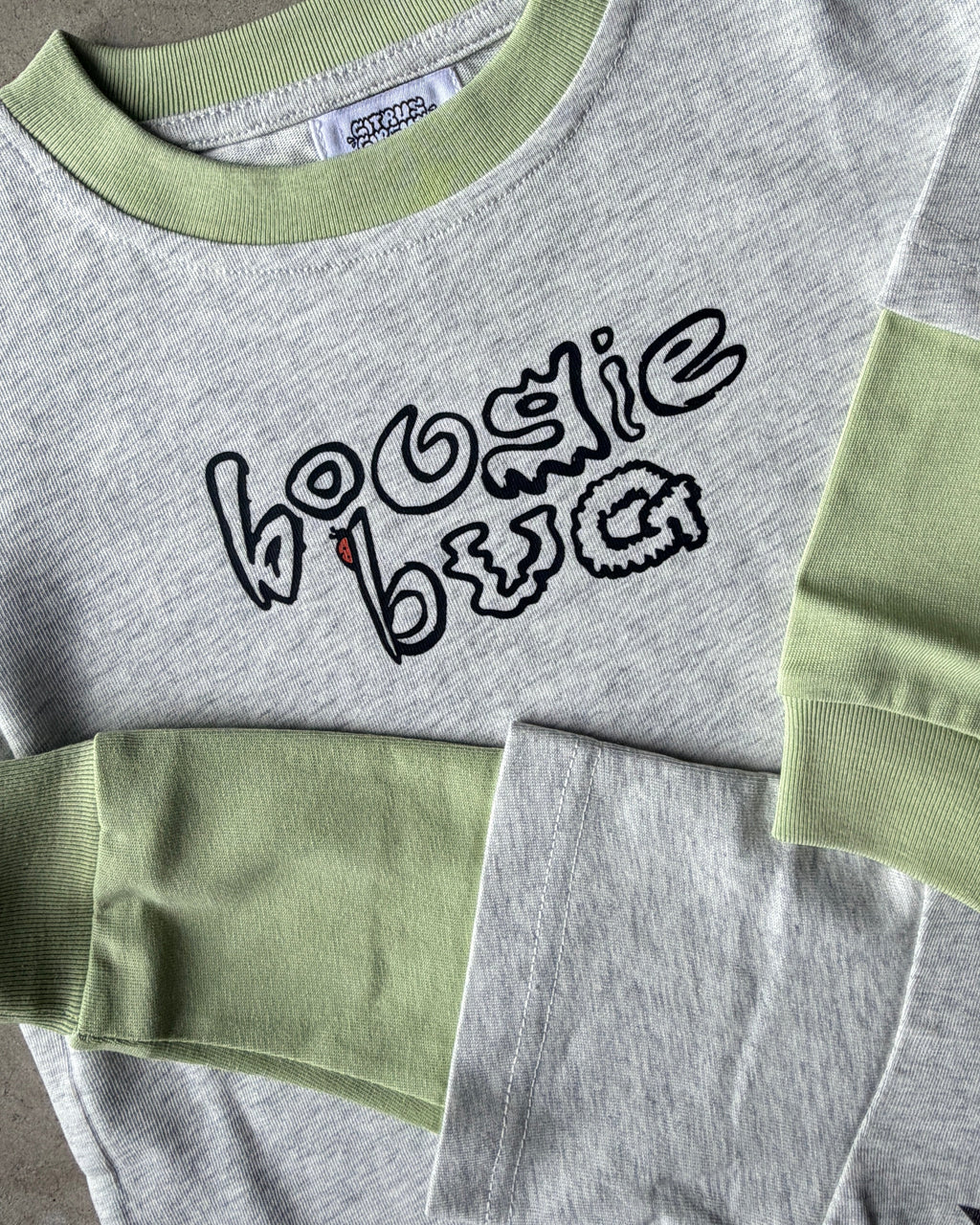 Gray t shirt with green sleeves and collar, featuring black text that reads 'boogie bug' on a concrete surface.