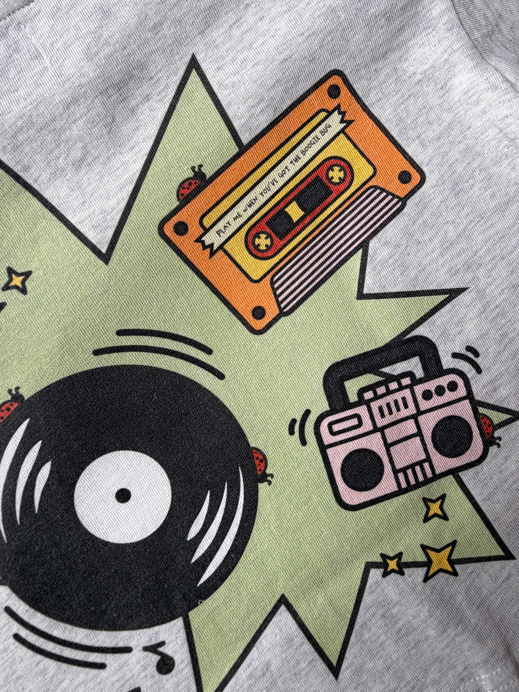 Gray t-shirt with a graphic design of a cassette tape, vinyl record, and boombox on a green starburst background with ladybugs.