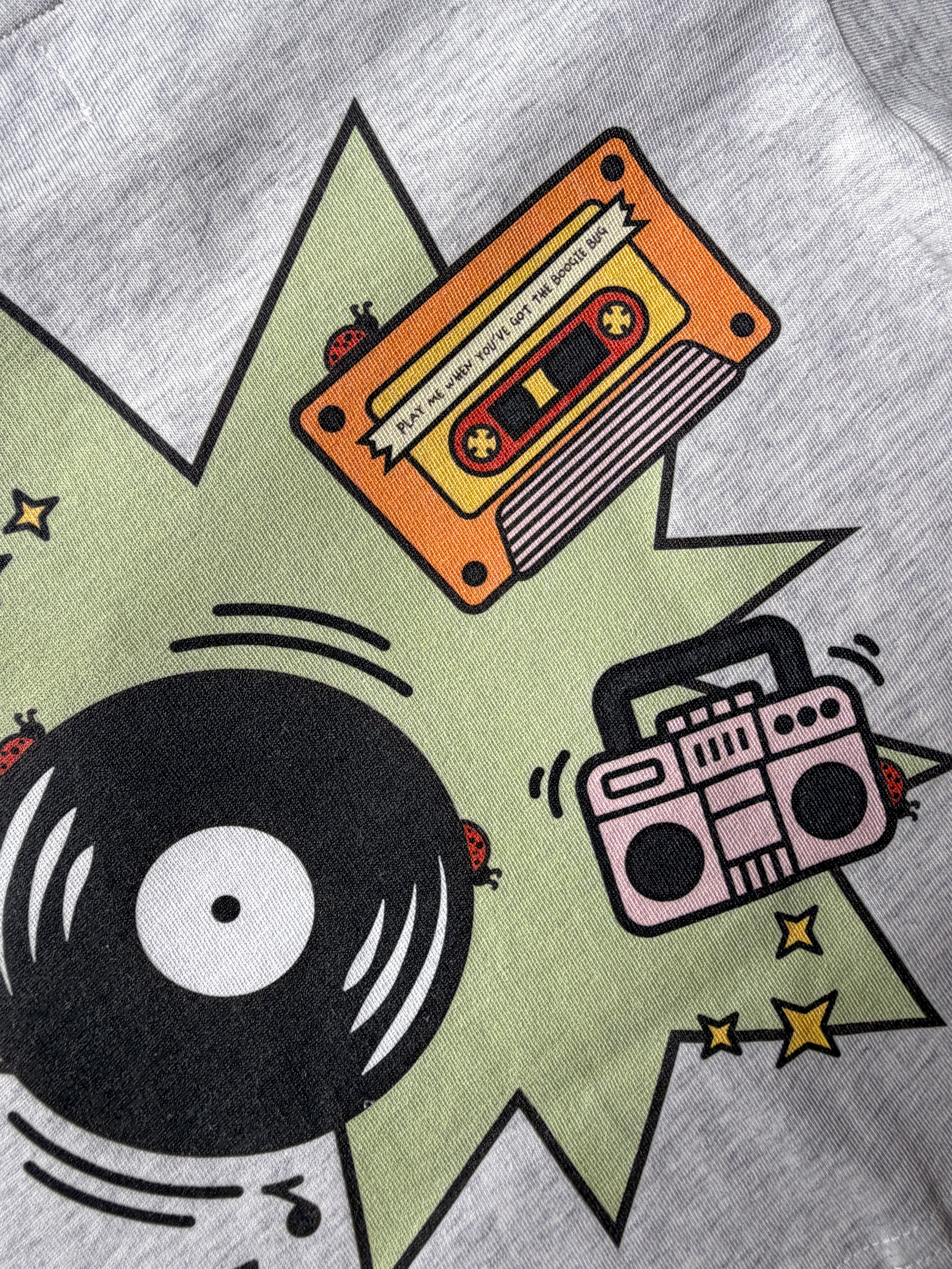 Gray t-shirt with a graphic design of a cassette tape, vinyl record, and boombox on a green starburst background with ladybugs.