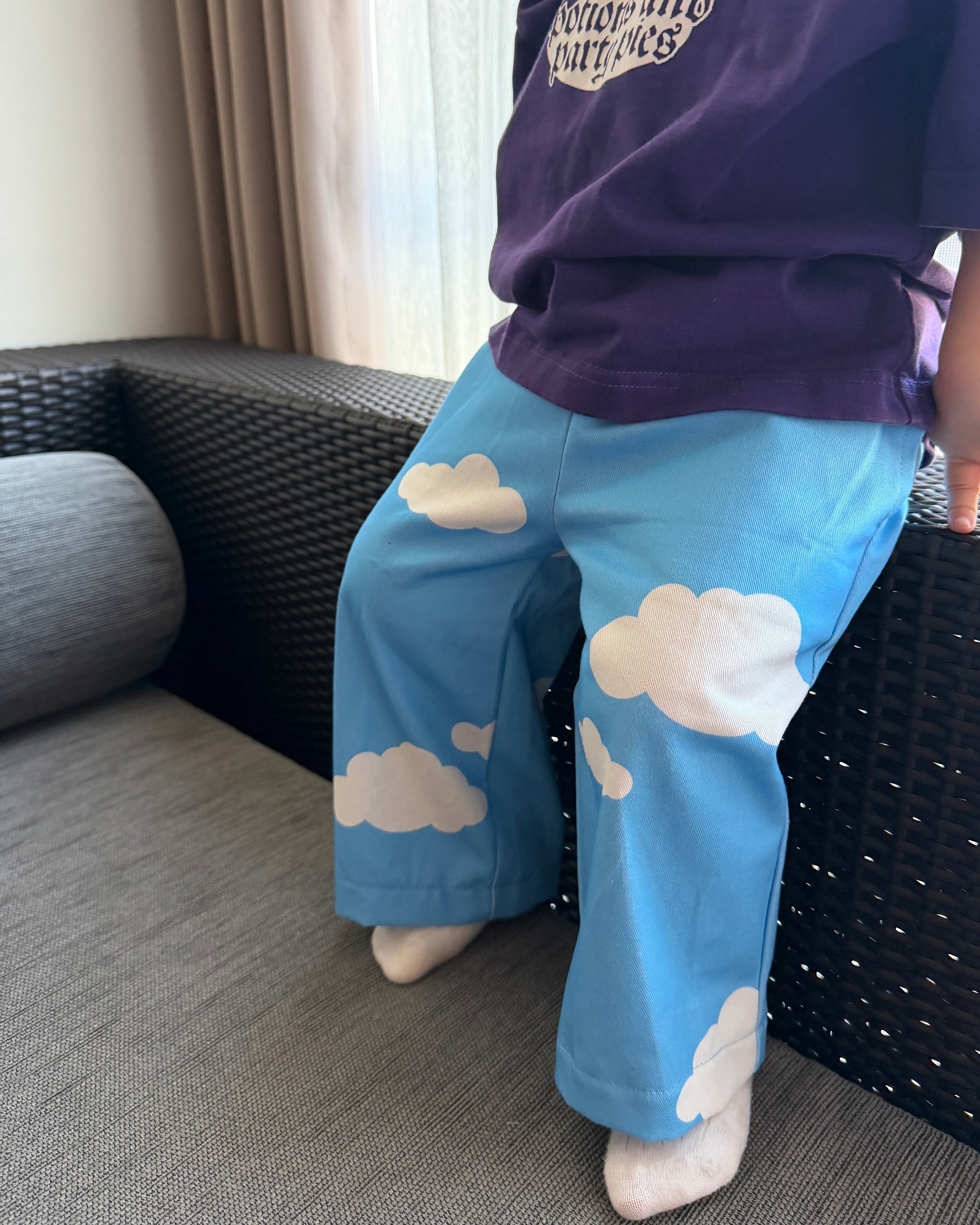Child wearing a purple shirt and blue pants with cloud patterns, sitting on a couch.