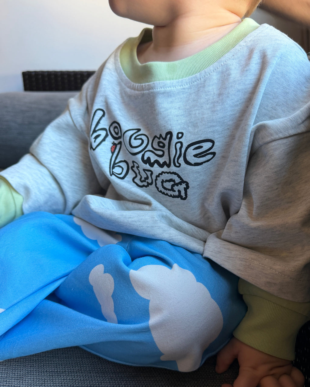 Child wearing a gray t shirt with 'Boogie Bug' text and blue pants with a white cloud design, sitting on a couch.