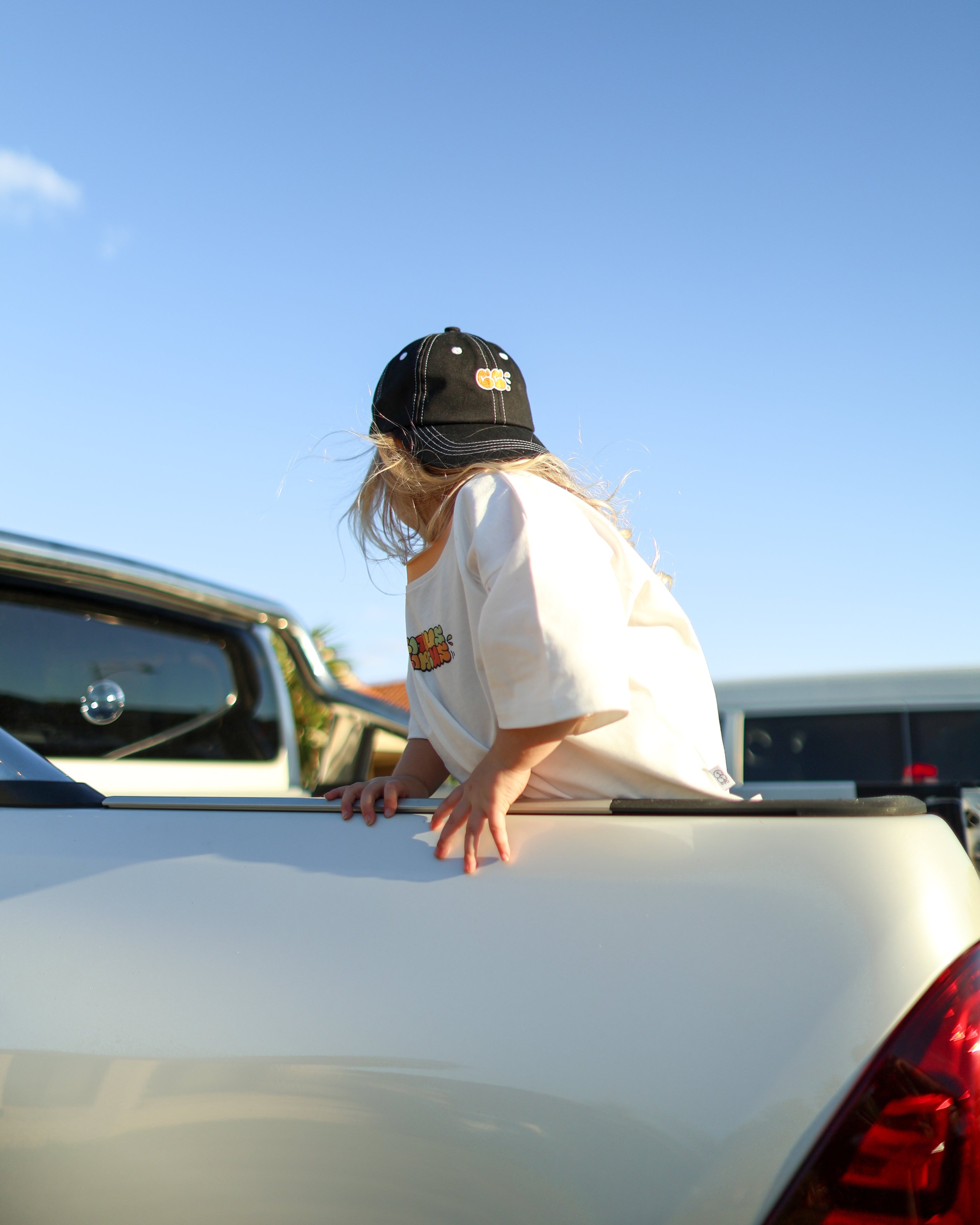Citrus Circus Starburst OG tee on child standing in Ute car tray - oversized kids white tee and black 6 panel cap facing backwards