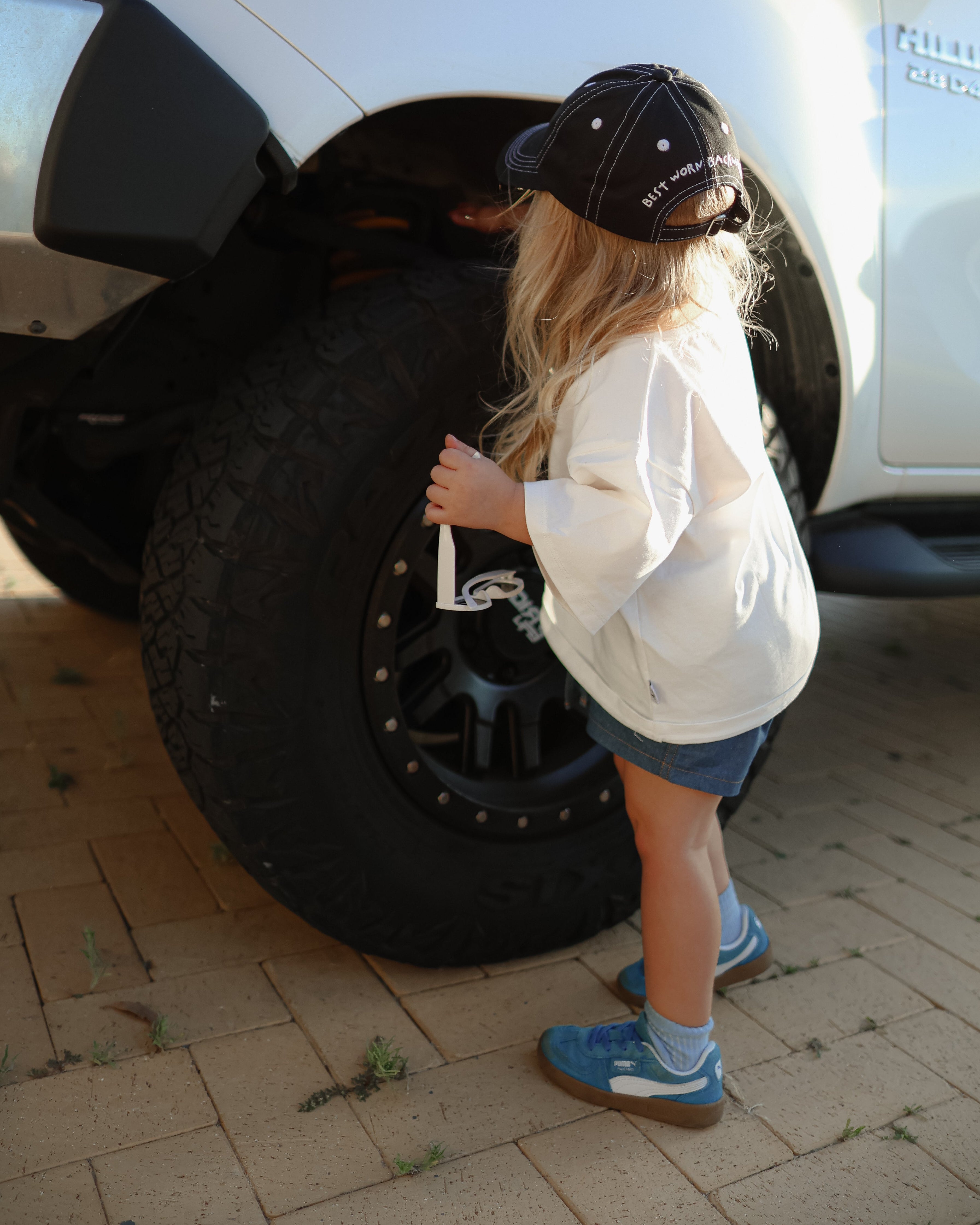 Citrus Circus Starburst OG Tee on child standing by car tyre - oversized kids white tee and navy cotton shorts facing backwards wearing black cap with white stitching and blue shoes