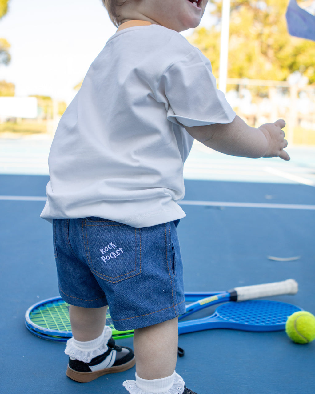 Citrus Circus Rock Pocket Shorts child at tennis court with tennis balls and rackets - oversized kids white tee and navy cotton shorts facing backwards