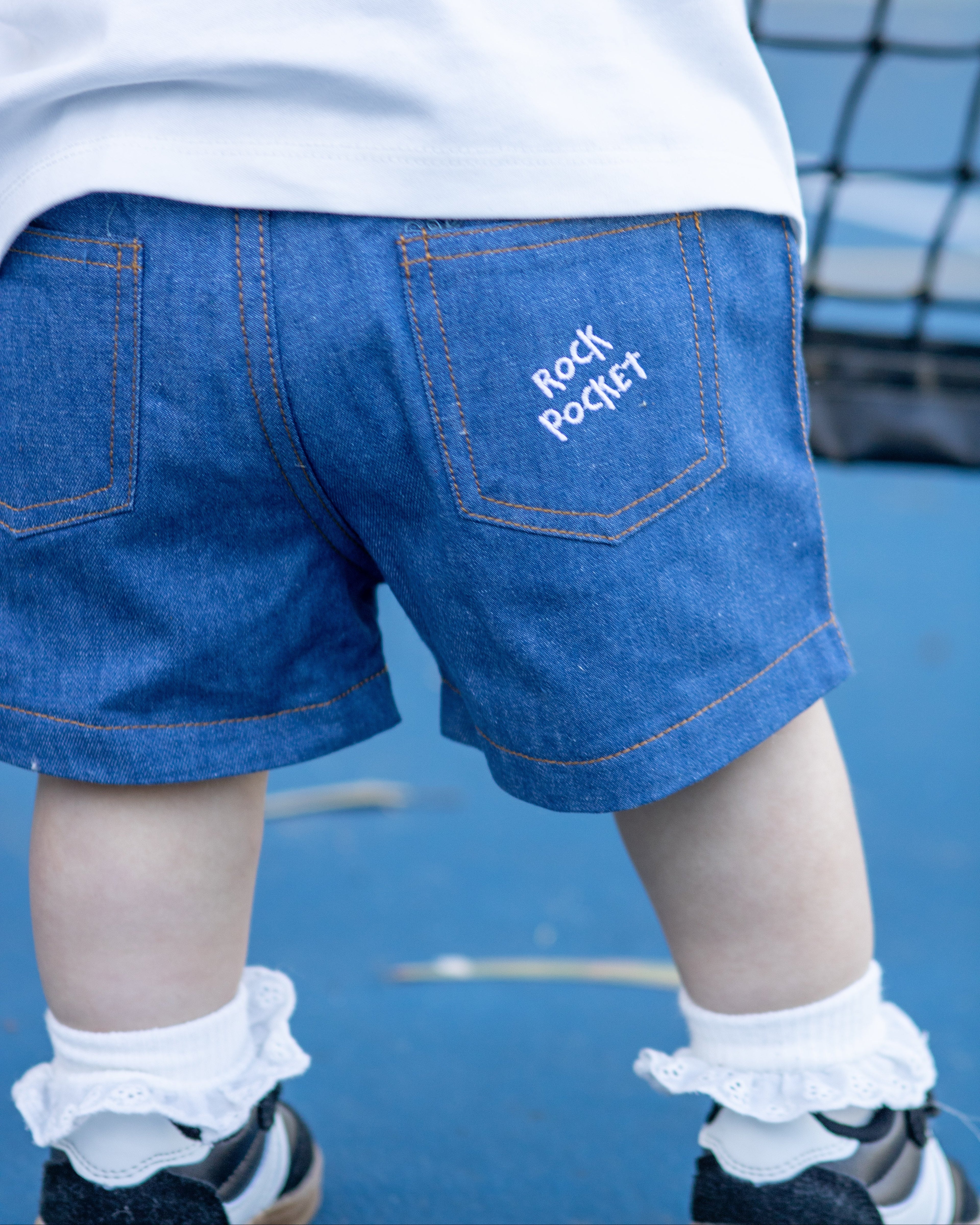 Citrus Circus Rock Pocket Shorts on child at tennis court - navy cotton shorts facing backwards with frilly socks and black shoes