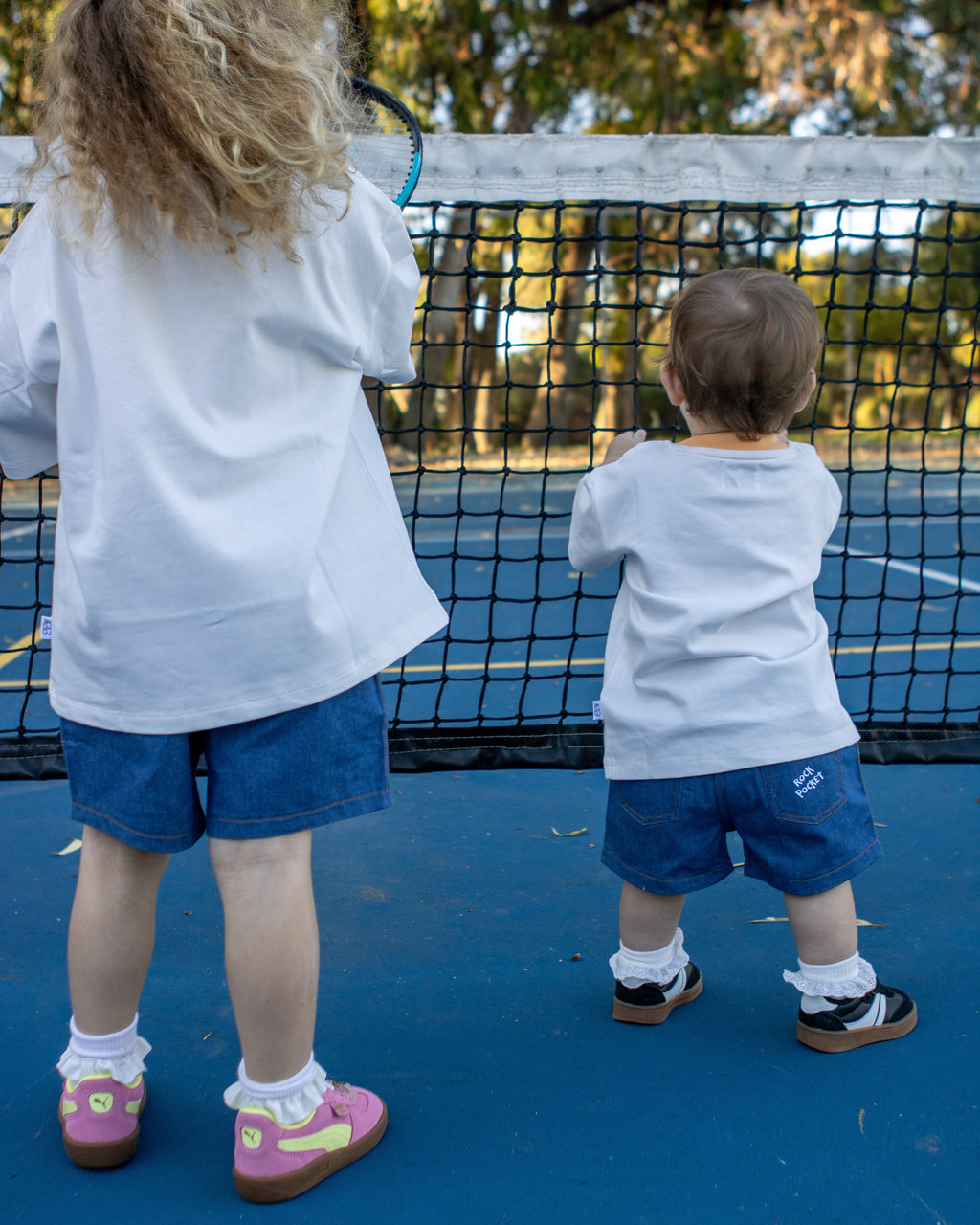 Citrus Circus Rock Pocket Shorts on two children at a tennis court - oversized kids white tee and navy cotton shorts facing backwards in frilly socks and sneakers