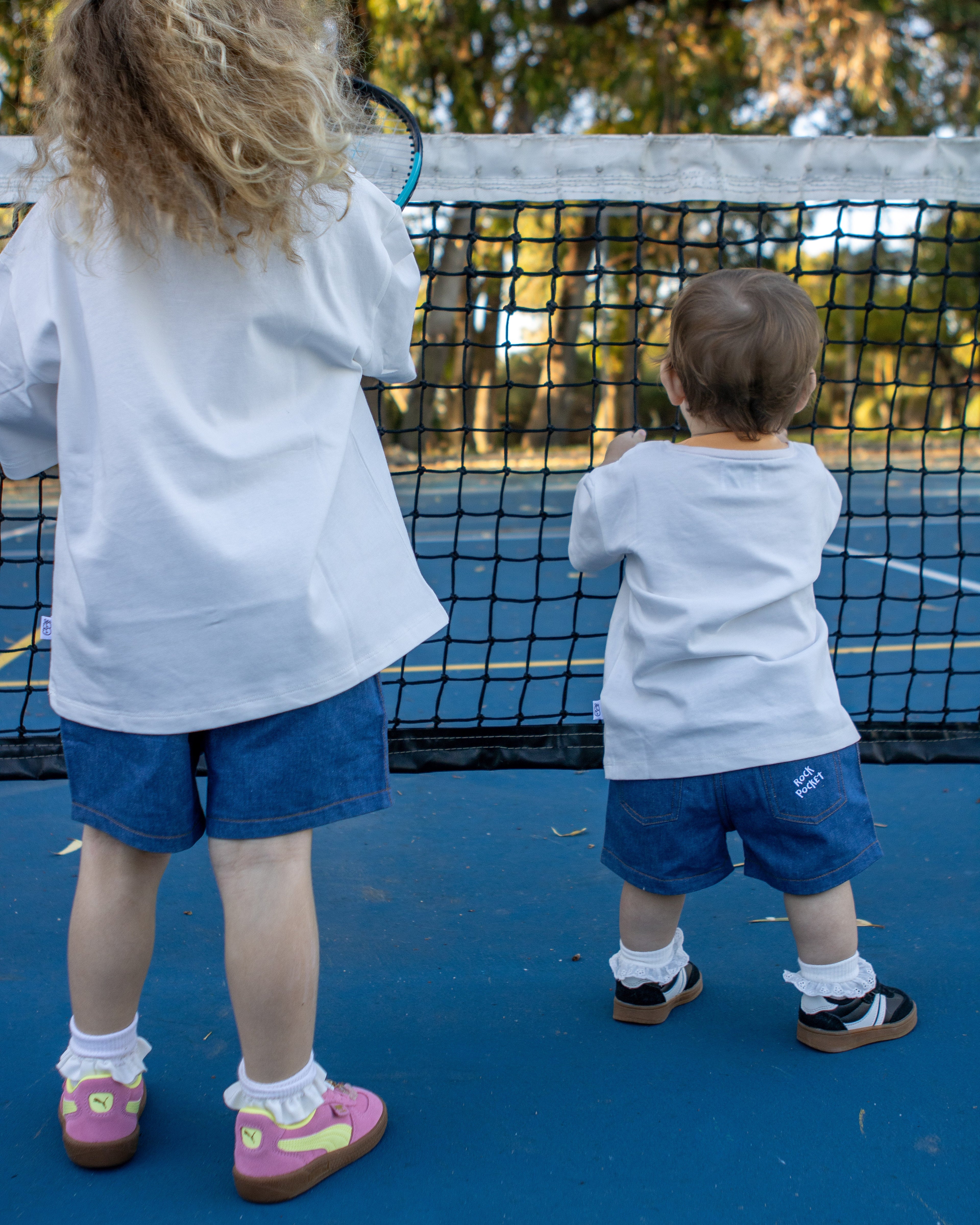 Citrus Circus Rock Pocket Shorts on two children at a tennis court - oversized kids white tee and navy cotton shorts facing backwards in frilly socks and sneakers