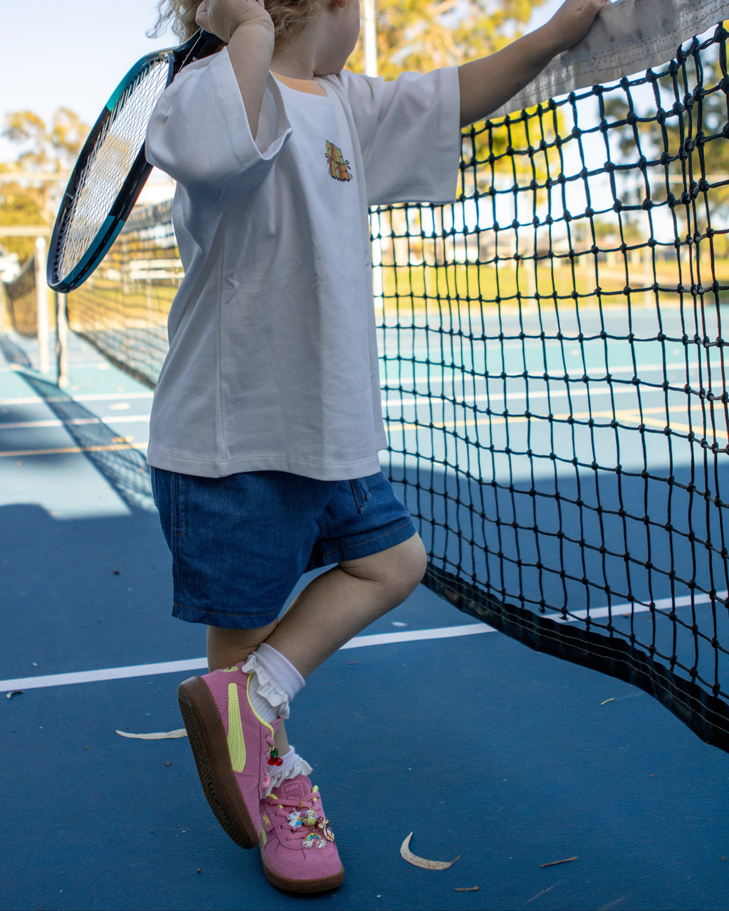 Citrus Circus Starburst OG Tee child at tennis court - oversized kids white tee and navy cotton shorts with pink shoes holding a tennis racket