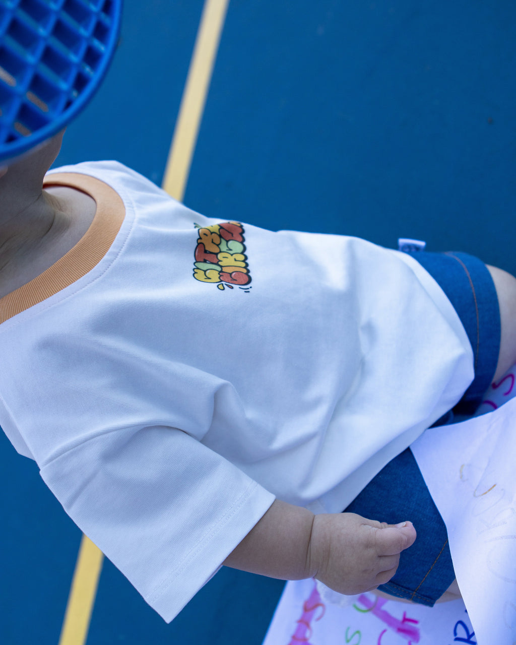 Citrus Circus Starburst OG tee on child at tennis court - oversized kids white tee with orange collar and navy cotton shorts facing sideways with tennis racket