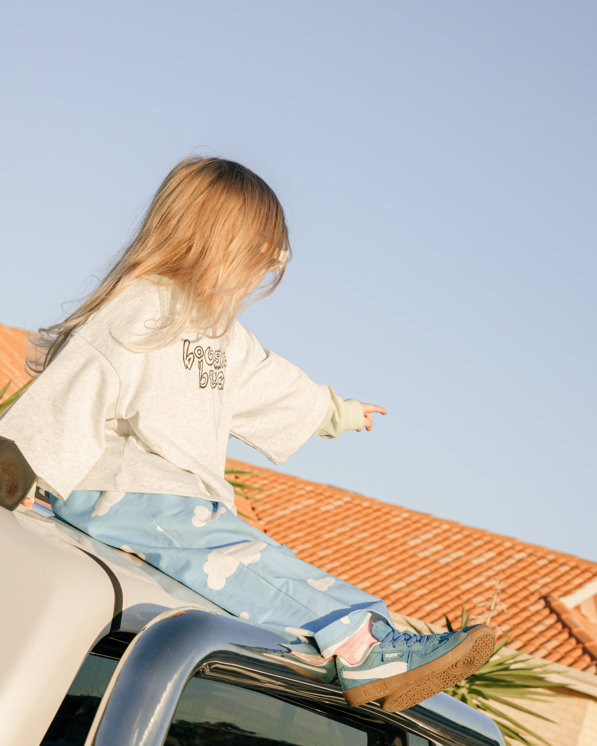 Child sitting on top of car in blue cloud pants and an oversized grey t shirt with lime green sleeves.