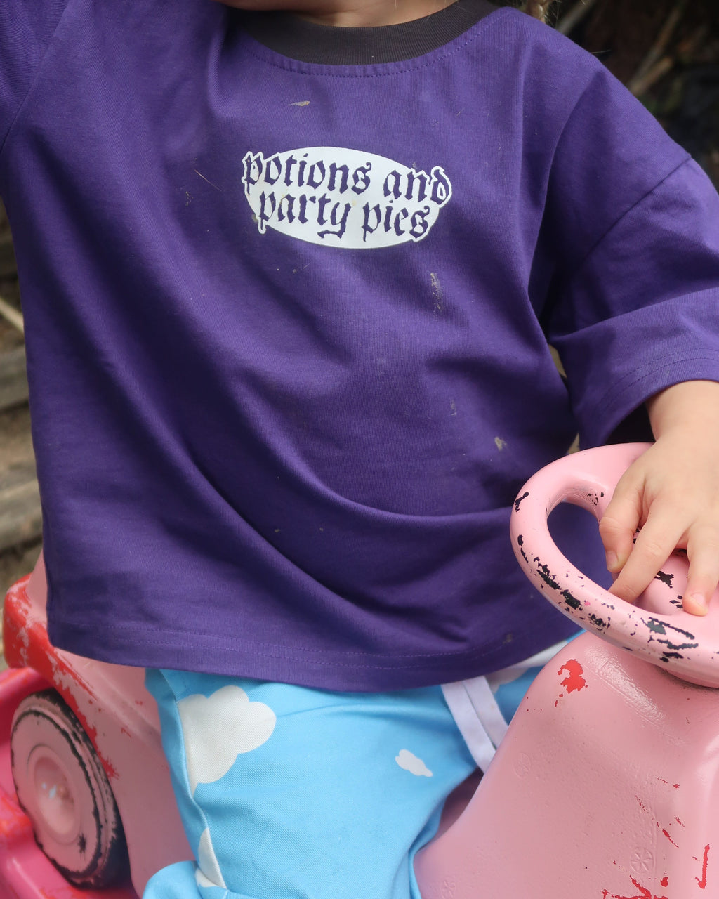 A close-up lifestyle shot of a child wearing a purple long-sleeve shirt with a white graphic print that reads 'Potions and Party Pies'. The child is sitting in a pink toy car outdoors.