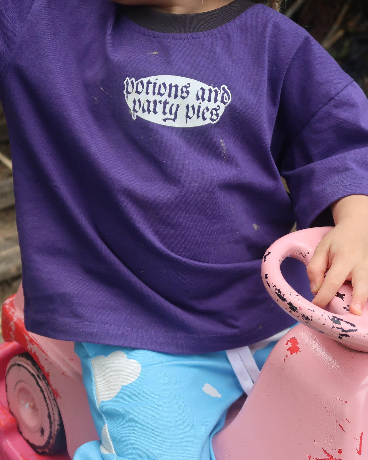 A close-up lifestyle shot of a child wearing a purple long-sleeve shirt with a white graphic print that reads 'Potions and Party Pies'. The child is sitting in a pink toy car outdoors.