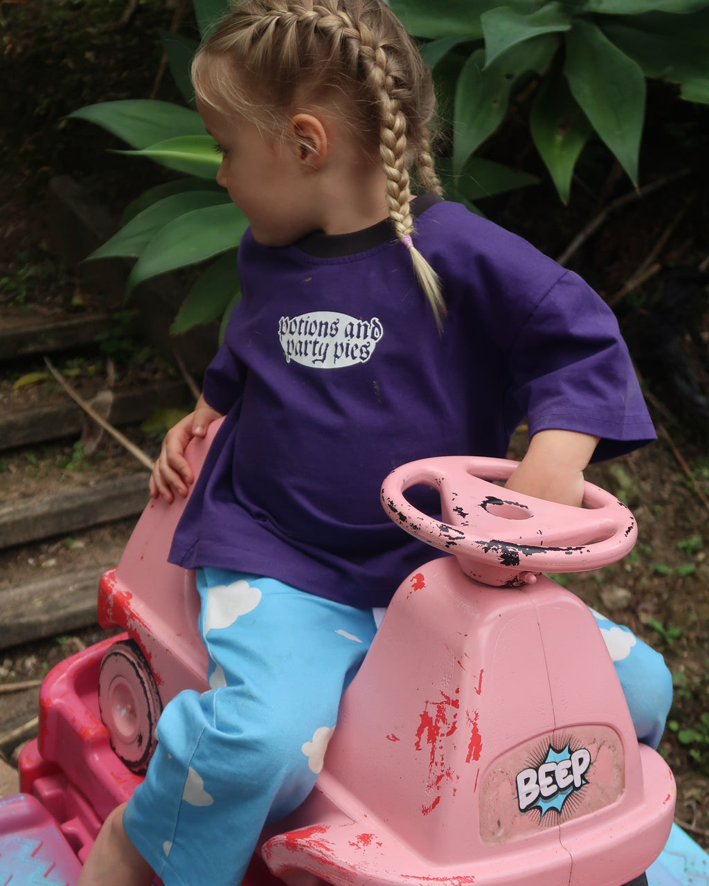 A young girl with braided hair sits in a pink ride-on toy car outdoors, wearing a purple t-shirt that reads 'Potions and Party Pies' and light blue cloud pants. The toy features a pink body with red splatter details.
