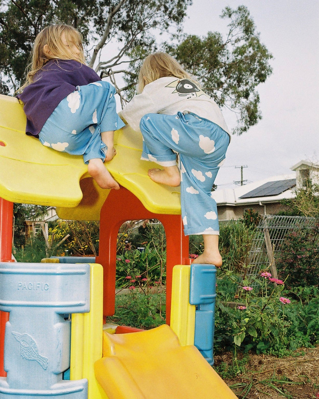 Two children in blue cloud pants sliding down a colorful playground slide on a sunny day.