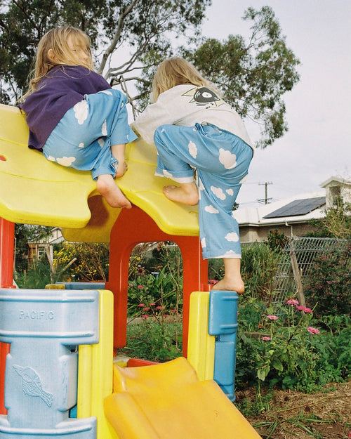 Two children in blue cloud pants sliding down a colorful playground slide on a sunny day.