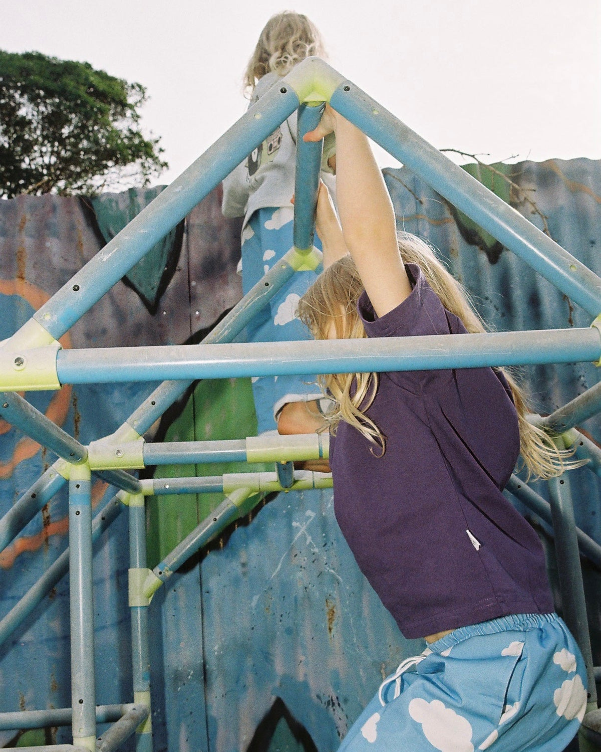 Two children climbing a metal playground structure with graffiti in the background. One wearing a purple shirt and both wearing blue cloud pants.