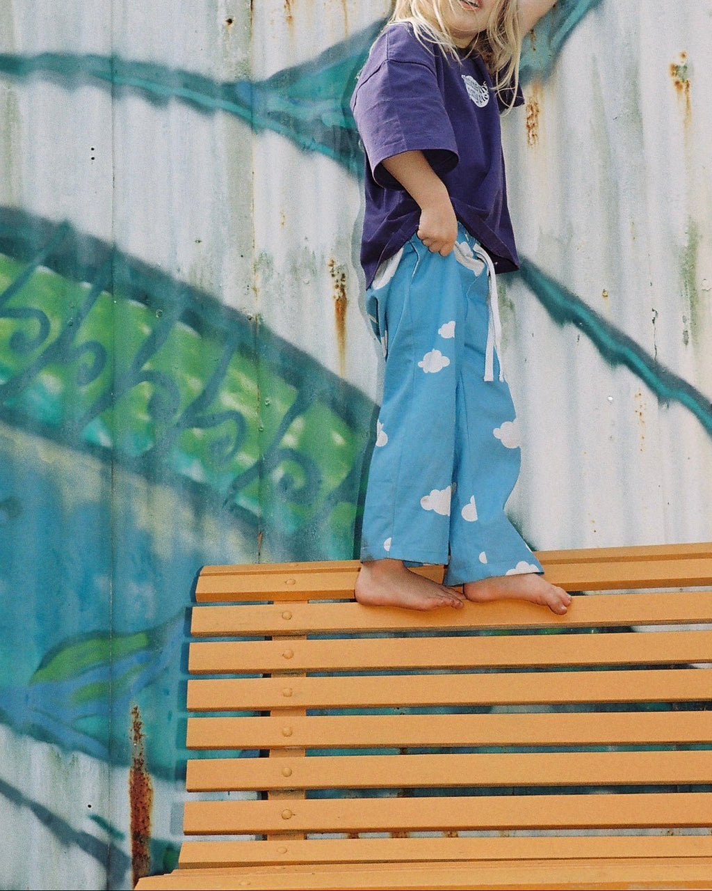 Child wearing a purple shirt and blue pants with cloud patterns on a wooden bench against a graffiti-covered wall.