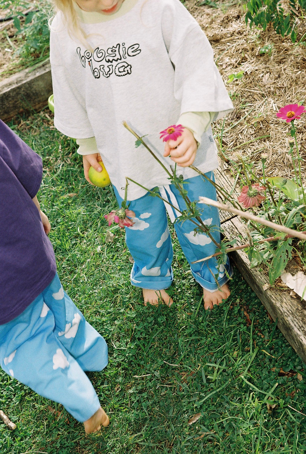 Two children in a garden, one holding a lemon and the other a stick with flowers, wearing matching outfits.